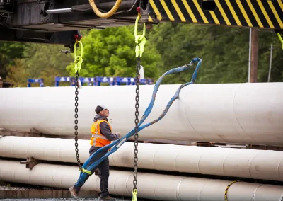 Industrial worker rigging large white coated pipes with a sling and overhead hoist chains.